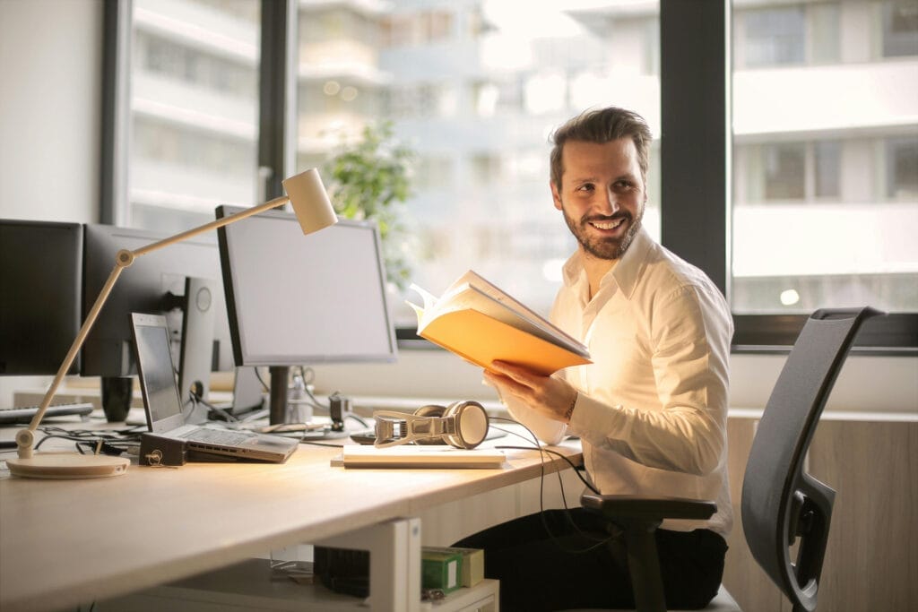 Smiling man reading book at desk in office.