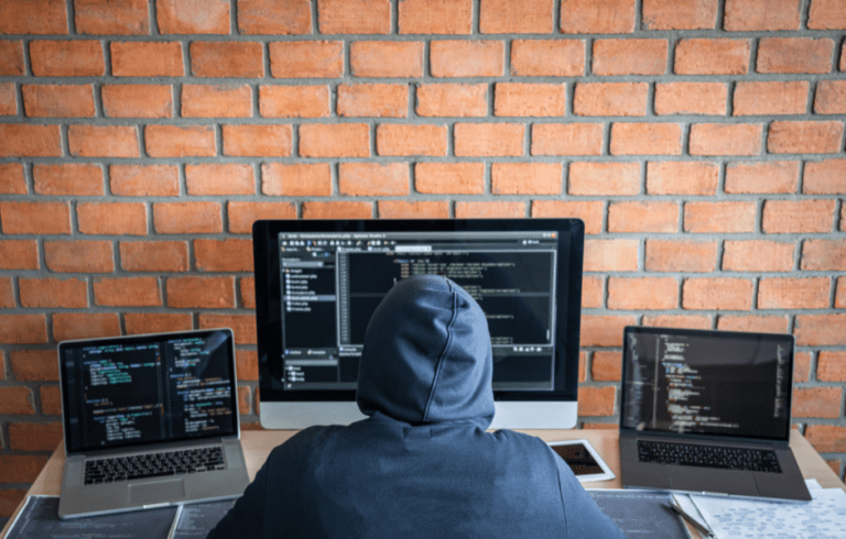 Hacker coding with multiple screens in brick-walled room.