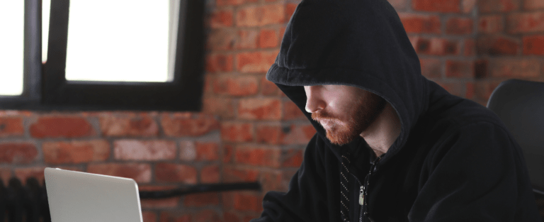 Hooded person working on laptop in dim room.